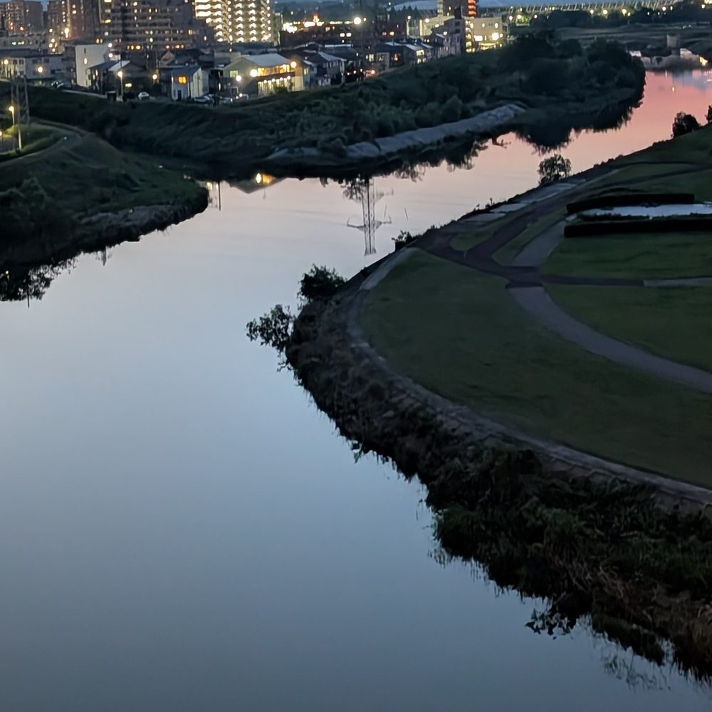 夕暮れの川と対岸の街並み、手前は公園の芝生と池が見える風景写真